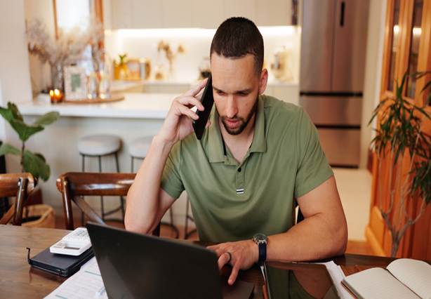 A man is on the phone while using a laptop at his dining table, appearing to work from home.
